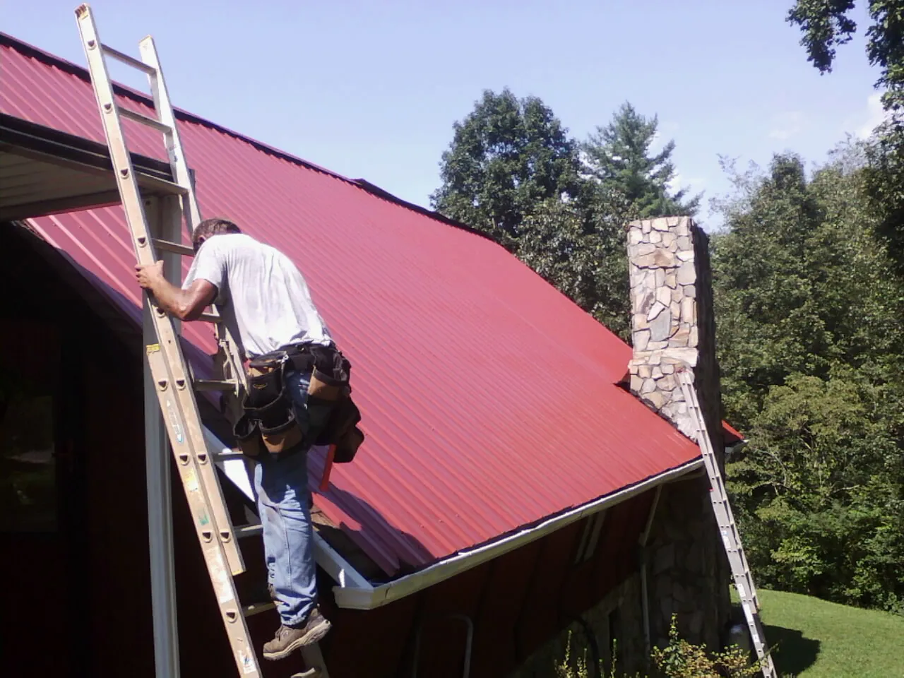 Roofer on a ladder beside a red metal roof and stone chimney — Soffit & Fascia Repair by Roofing Brothers in Prior Lake